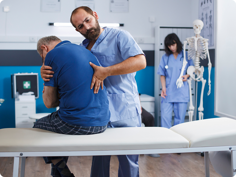 Doctor assisting an elderly patient with back mobility in an exam room.