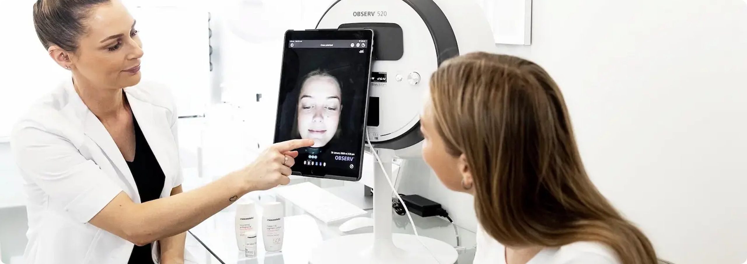 A clinician showing a female patient a digital facial scan on a tablet connected to a clinical imaging device.