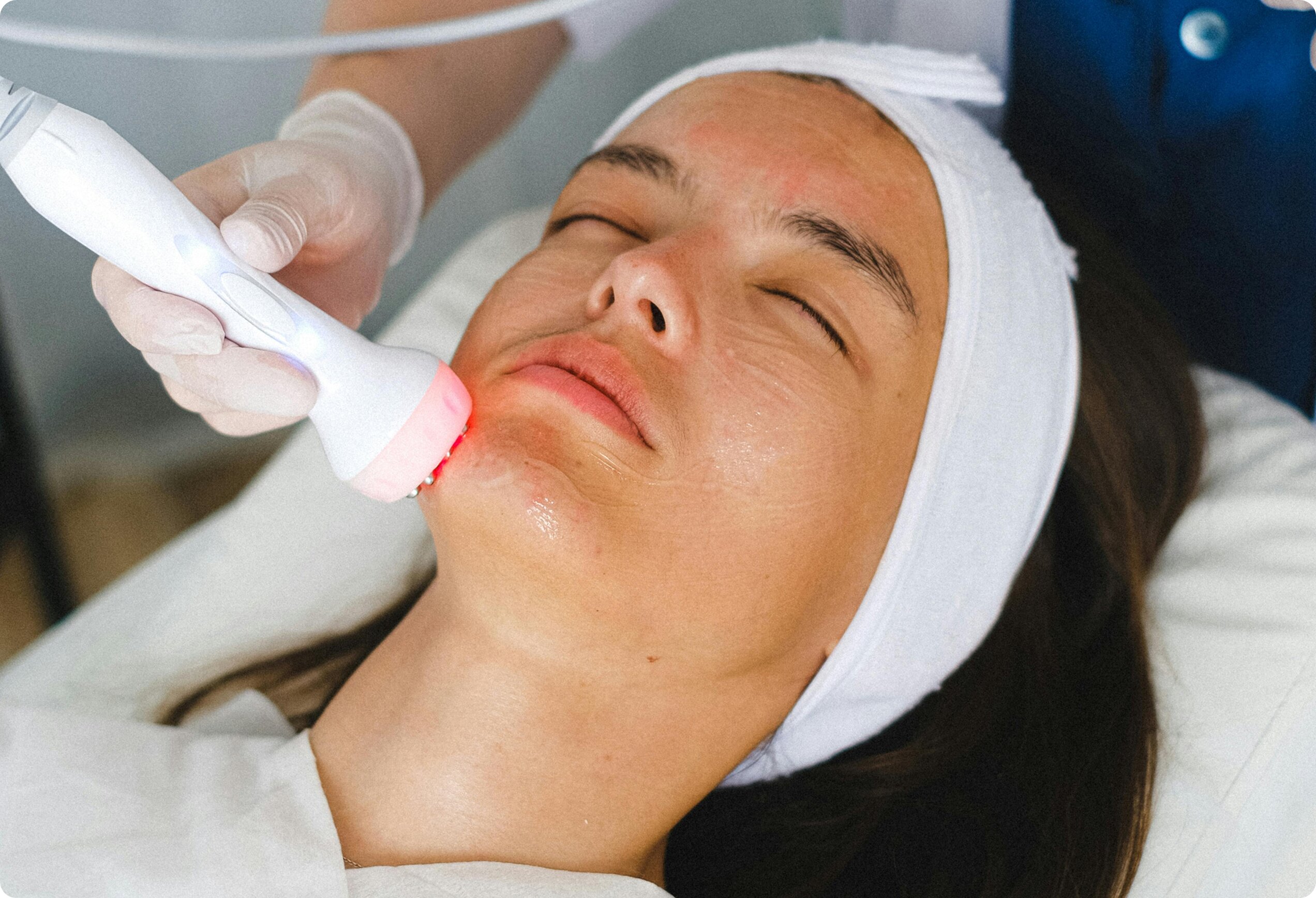 A close-up of a female patient receiving a red-light therapy facial treatment from a medical professional wearing gloves.