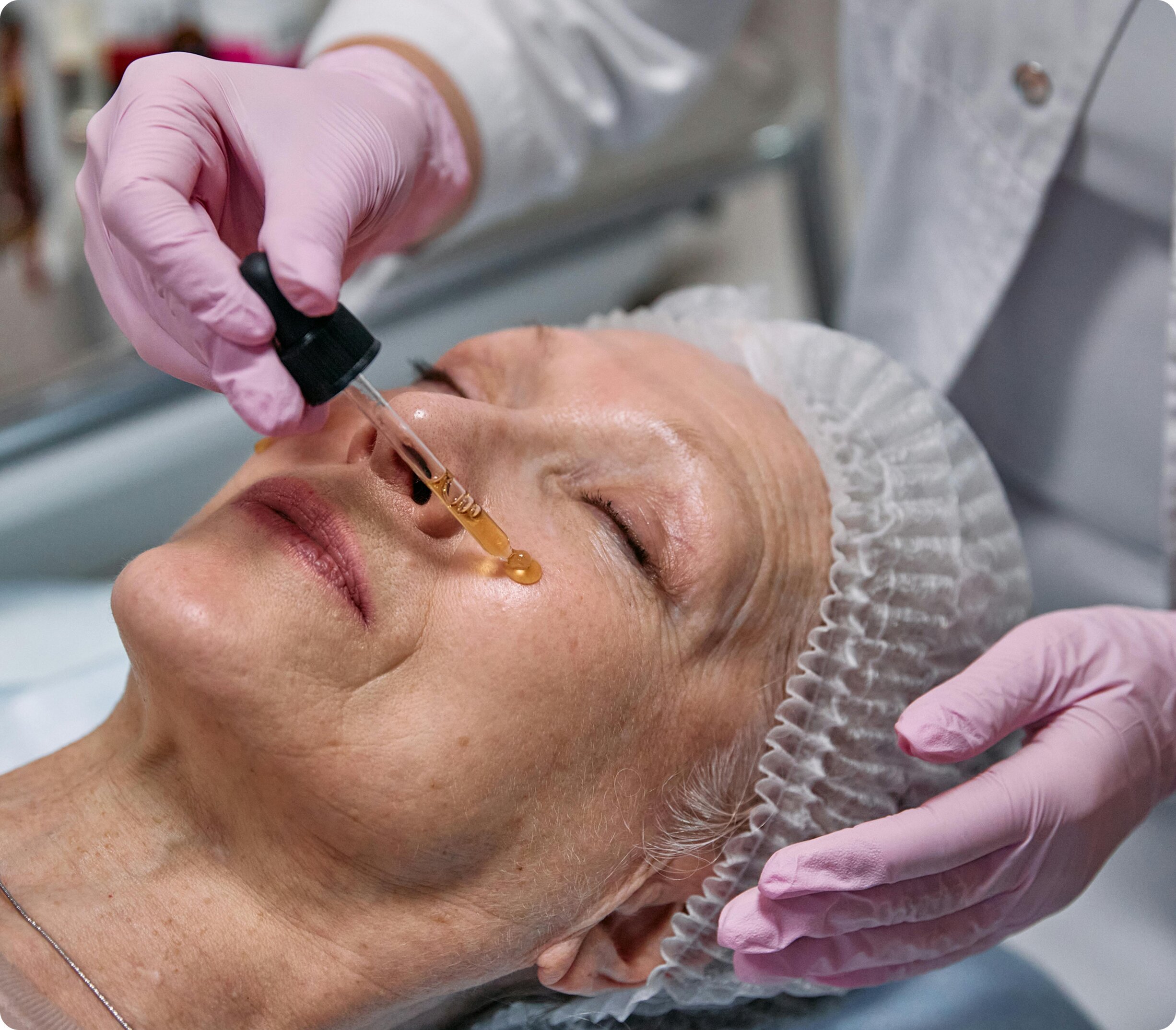 An older female patient resting comfortably while a medical professional in pink gloves applies an anti-aging serum to her cheek using a glass dropper.