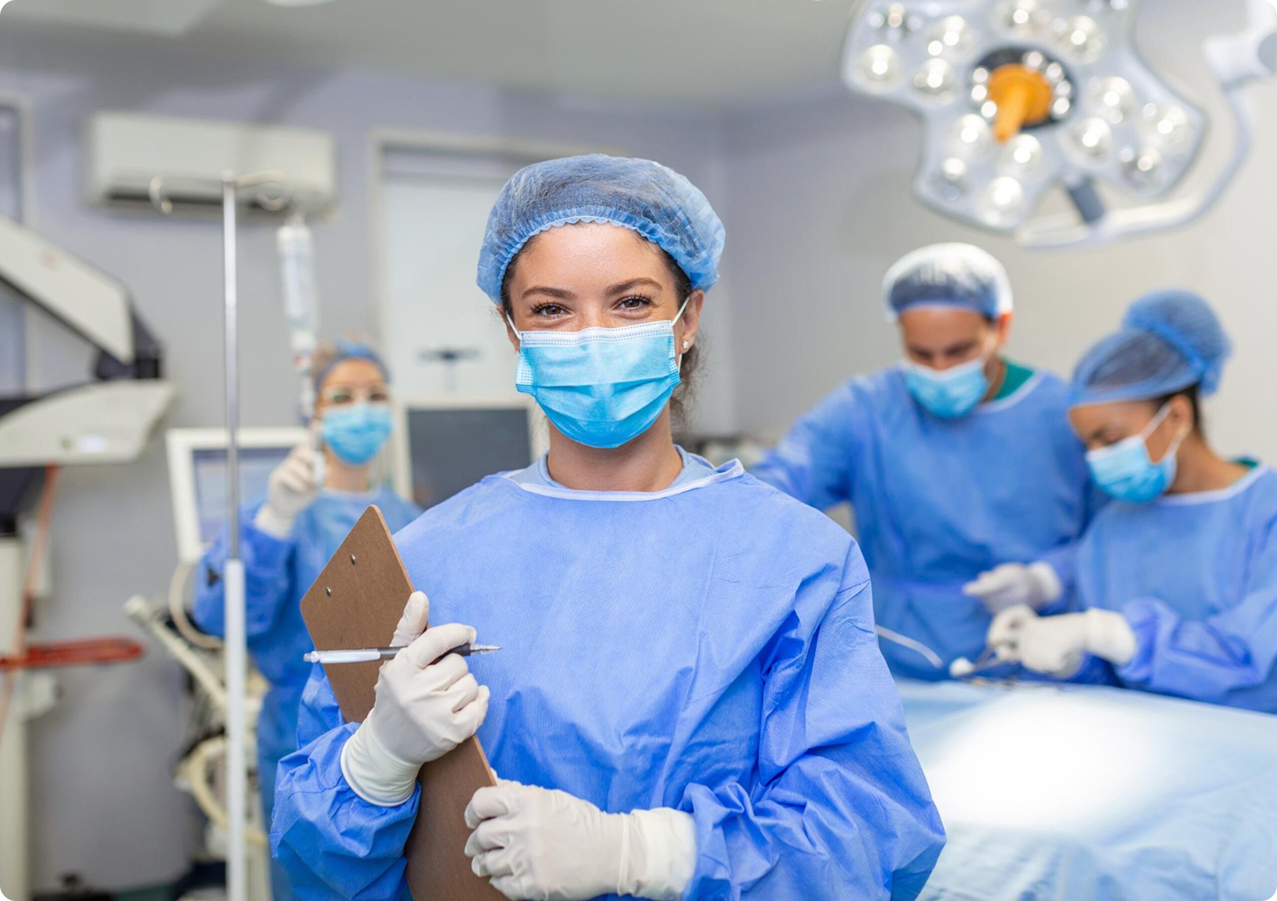 A smiling female medical professional in blue scrubs and a surgical mask holding a clipboard in a brightly lit operating room, with a surgical team working in the background.