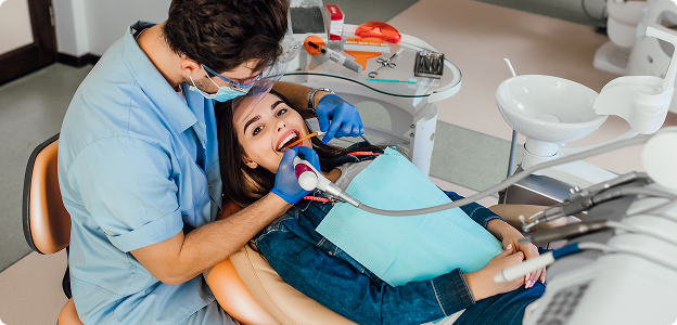 A dentist in blue scrubs and a face shield performing a procedure on a smiling female patient in a modern dental chair.