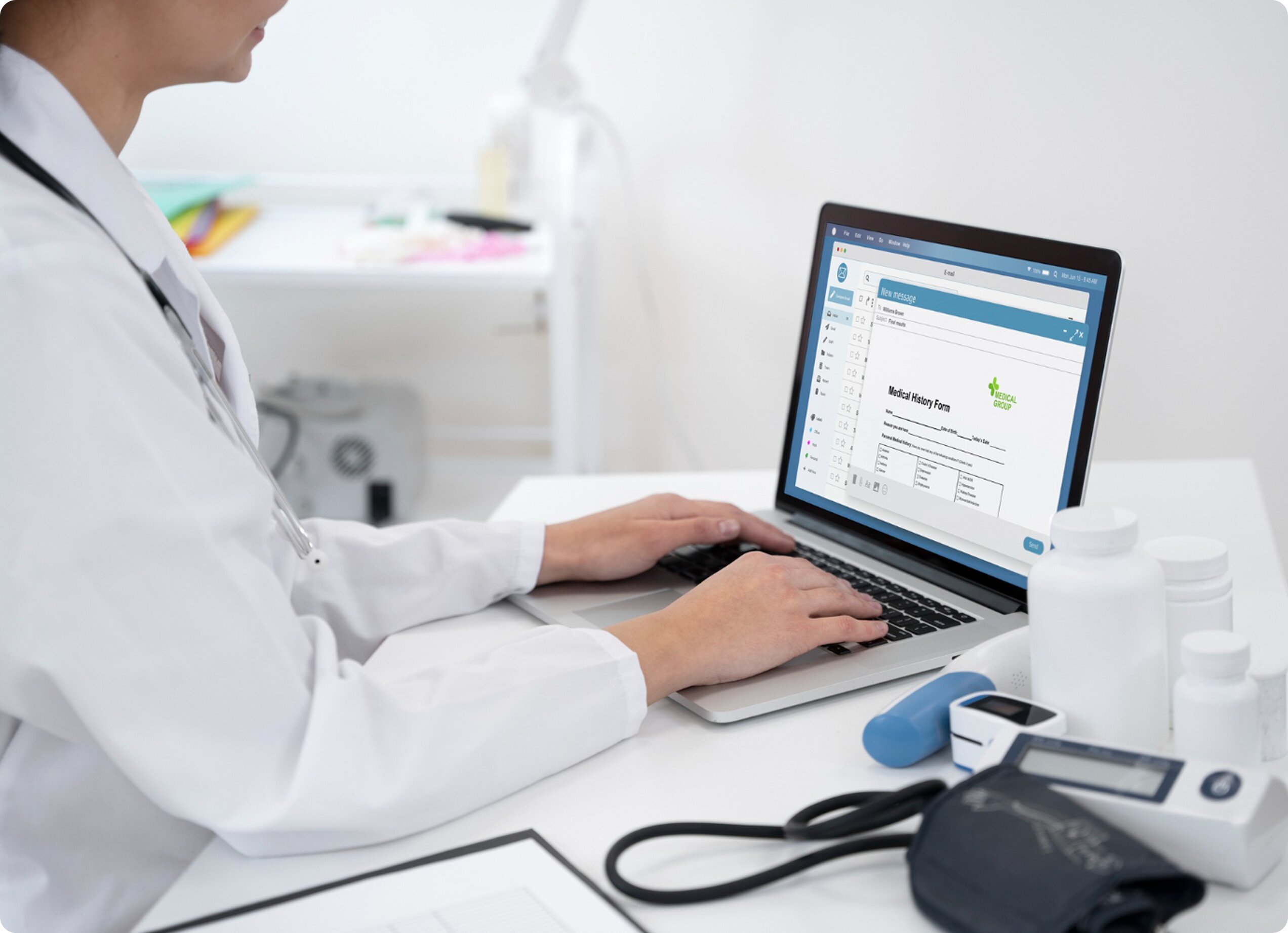 A doctor in a white lab coat typing on a laptop displaying a medical history form, with a blood pressure cuff and medical supplies on the desk.