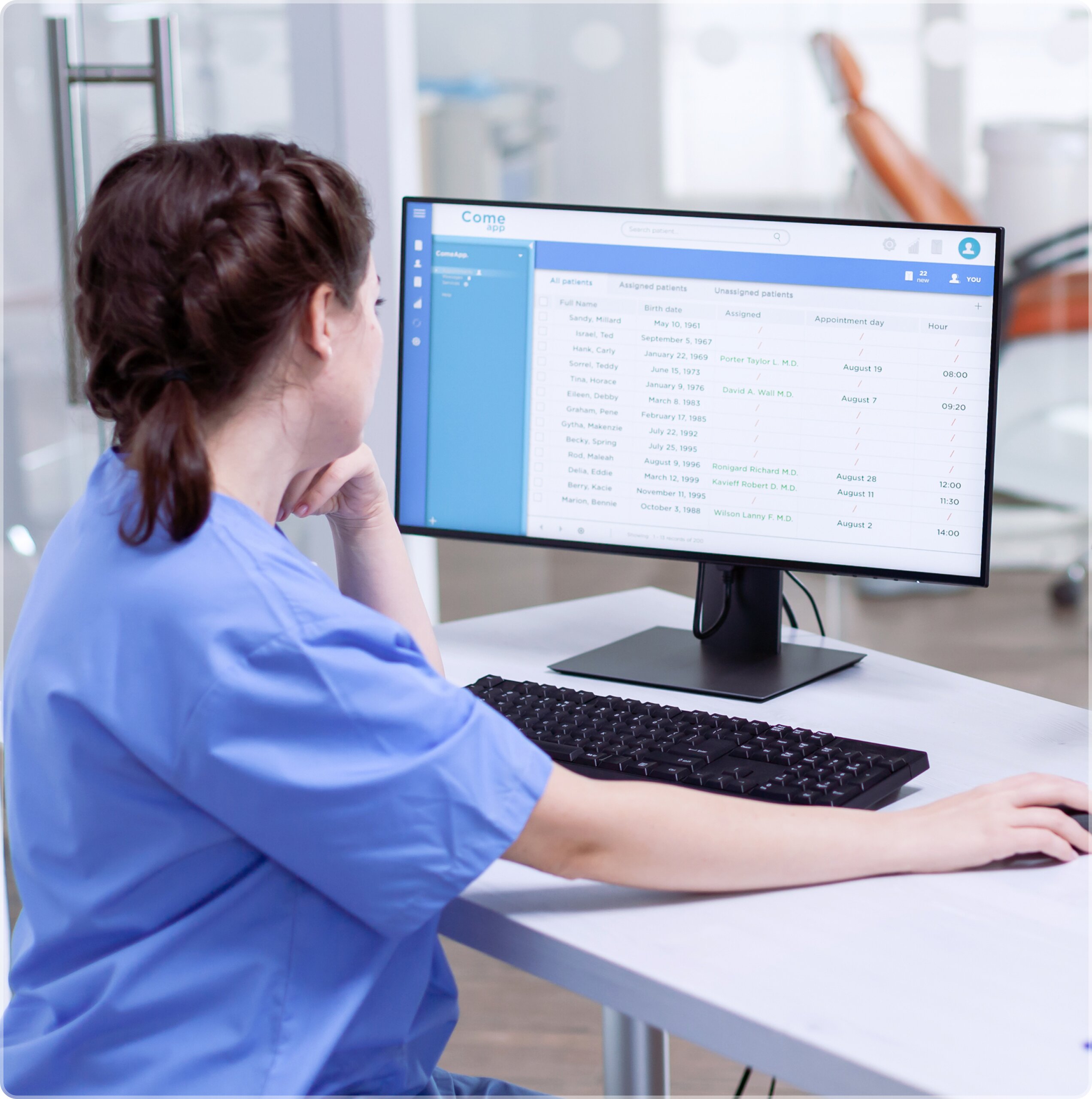 A medical professional in blue scrubs sitting at a desk, reviewing a patient appointment and management dashboard on a computer monitor.