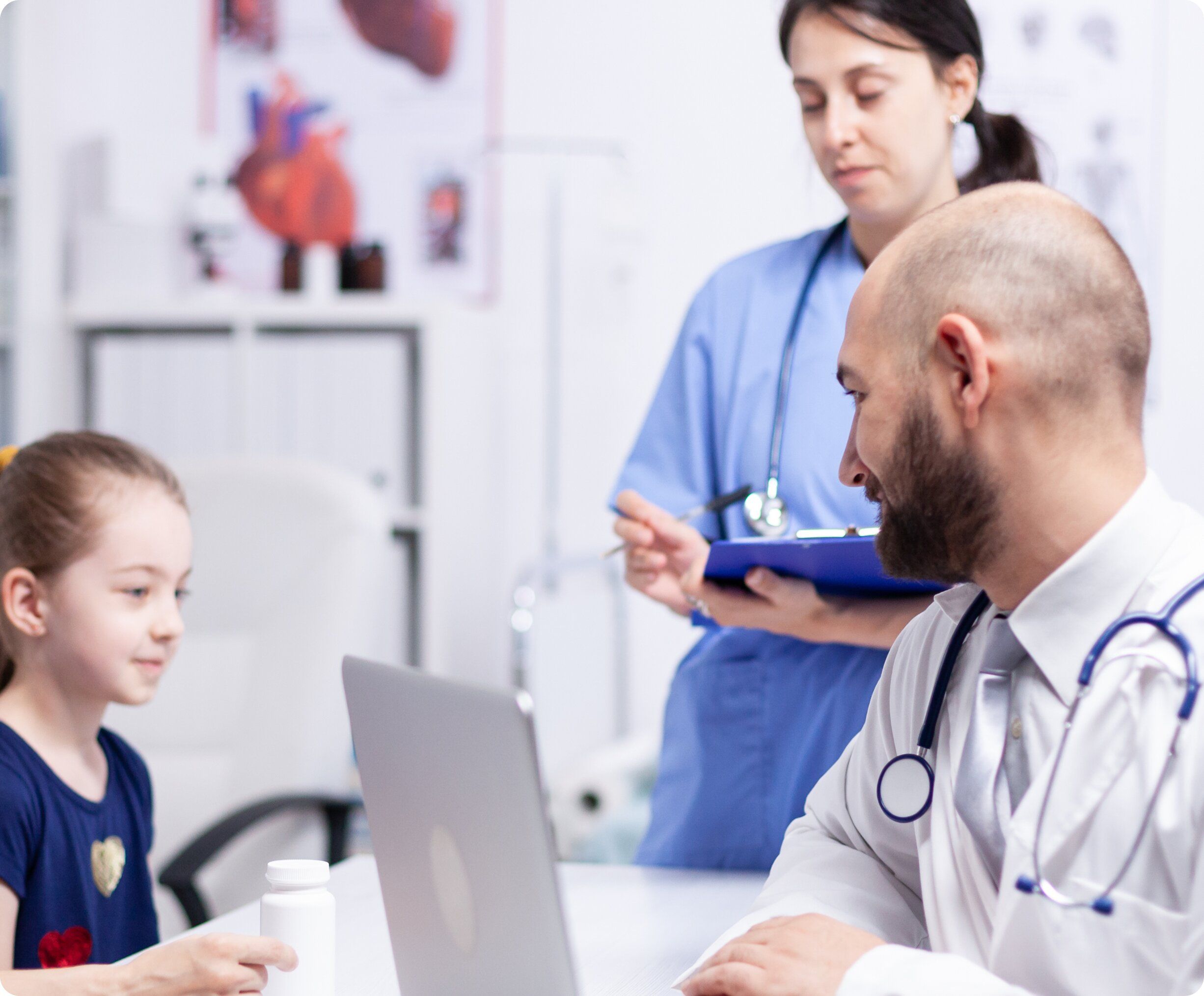 A male doctor in a white coat smiling and consulting with a young female patient at a desk, while a nurse in blue scrubs stands behind them taking notes on a clipboard.