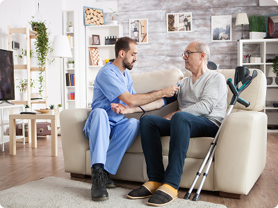 A male nurse in blue scrubs checking the blood pressure of an elderly man sitting on a sofa with crutches nearby in a home setting.