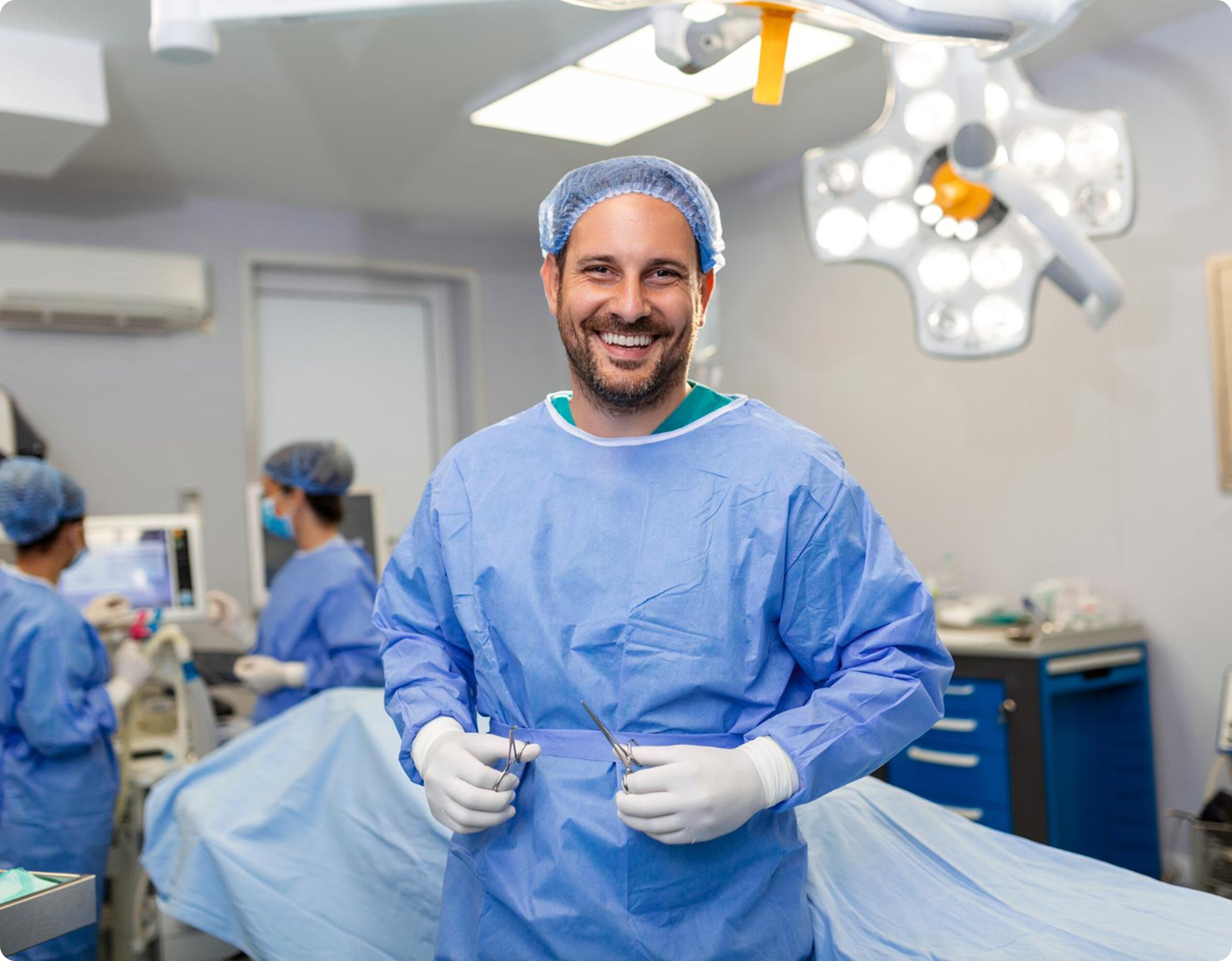 A smiling male plastic surgeon in blue scrubs, gloves, and a hairnet standing in a brightly lit operating room.