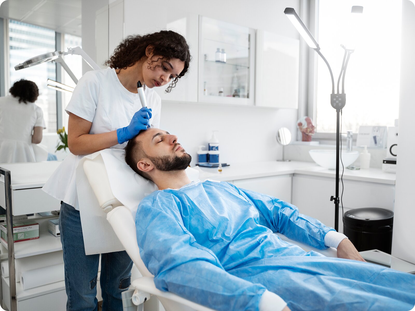 A male patient reclining in a treatment chair receiving a scalp or facial treatment from a female practitioner in blue gloves.