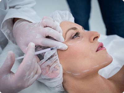 A medical professional in white gloves administering a dermal filler injection to the cheek area of a patient in a clinical setting.