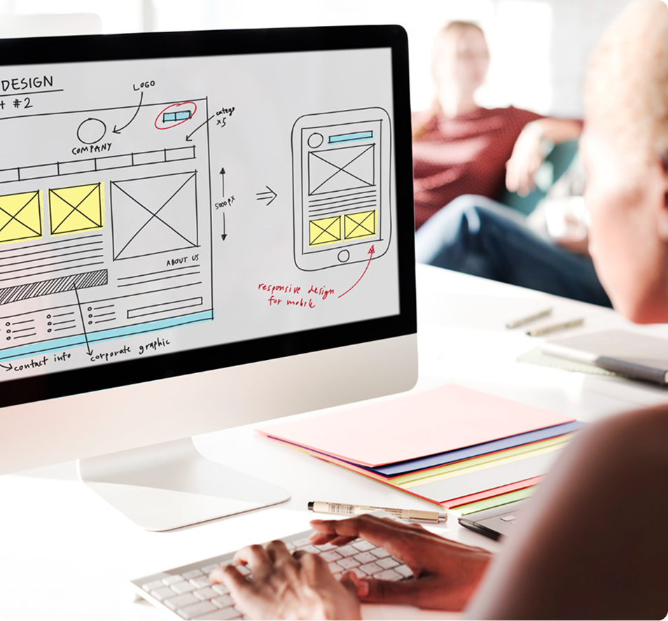 A web designer working on a desktop monitor showing a wireframe sketch of a medical website being converted into a mobile-responsive layout.