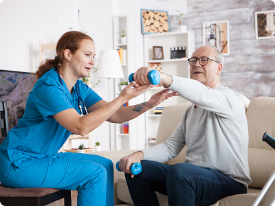A female physical therapist in blue scrubs assisting an elderly male patient with dumbbell exercises during a home or clinic rehab session.