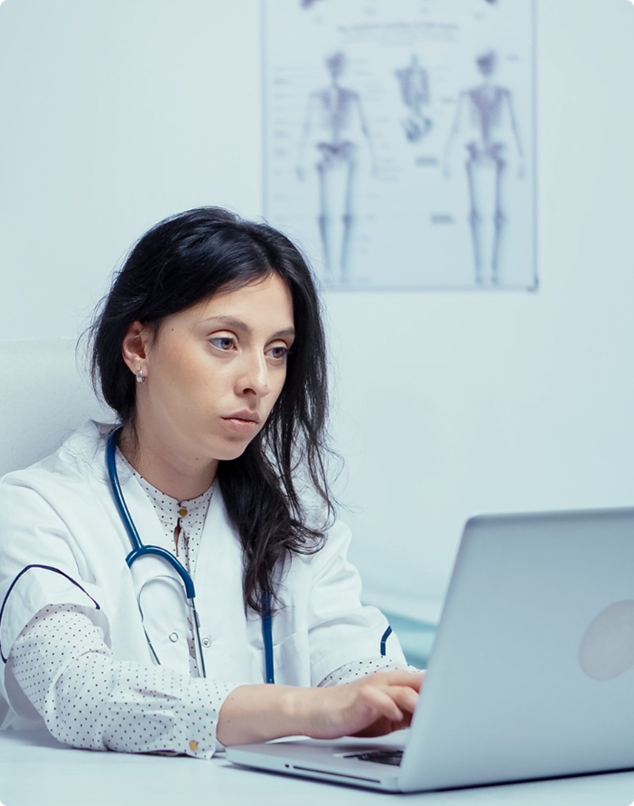 A female doctor in a white coat sitting at a clinic desk, typing on a silver laptop