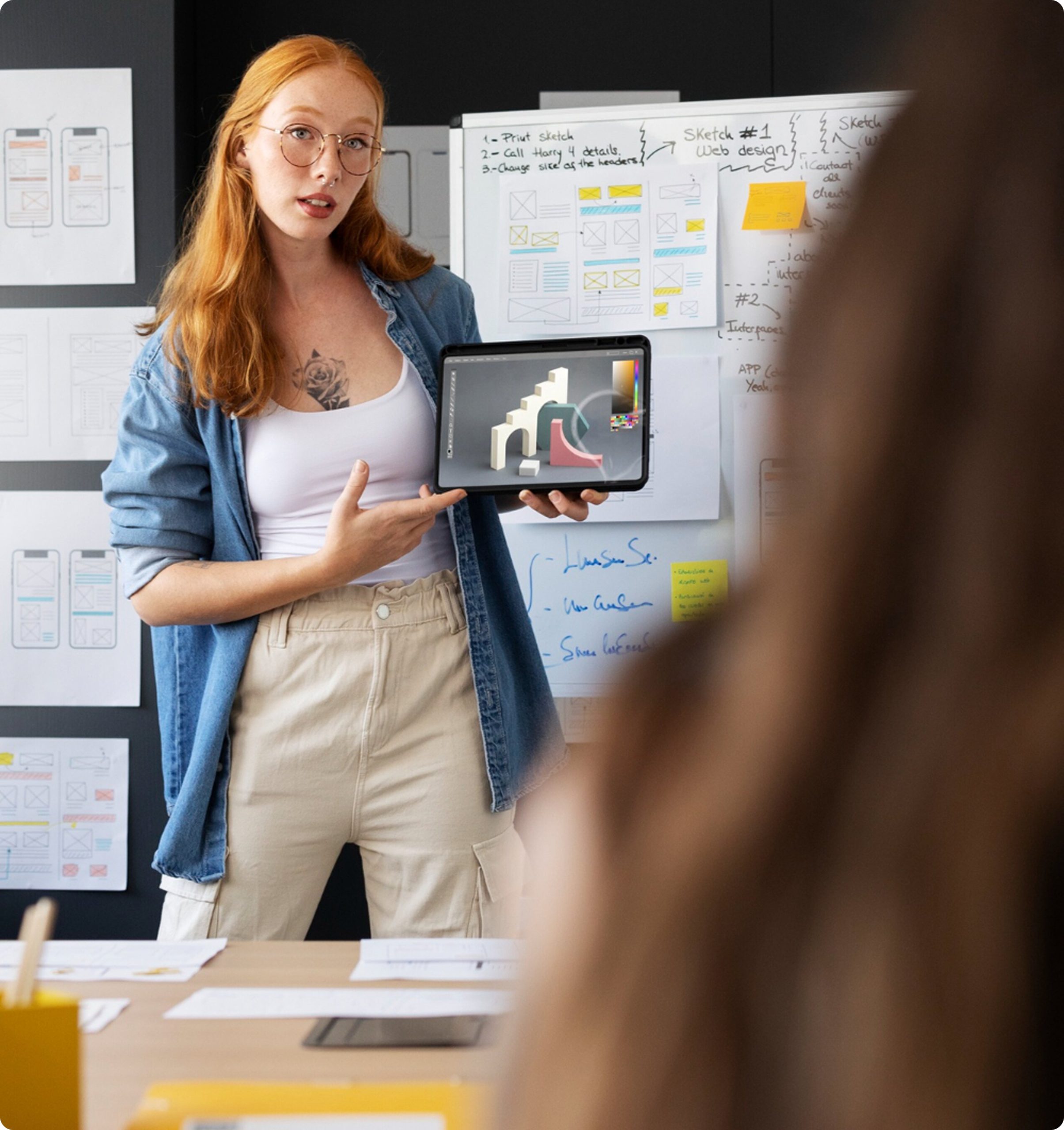 A digital marketing strategist standing in a modern office, presenting a design concept on a tablet with wireframes and a whiteboard in the background.